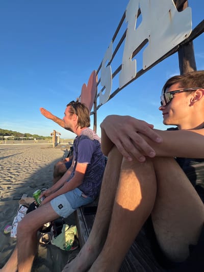 The image shows two men sitting on a bench on a sandy beach, likely in Pistula, Montenegro. The man on the right is wearing sunglasses and a dark t-shirt, with his arms crossed. The man on the left is wearing a purple t-shirt and blue shorts, holding a green can of beer and appears to be gesturing with his right hand. Scattered on the sand near the bench are various items including shoes, clothing, and another can. In the background, there are some trees and a clear blue sky. A white sign with large, cut-out letters is visible above the men, with a partial pink heart shape attached to the structure. The sun is low in the sky, casting long shadows and illuminating the scene with warm light.