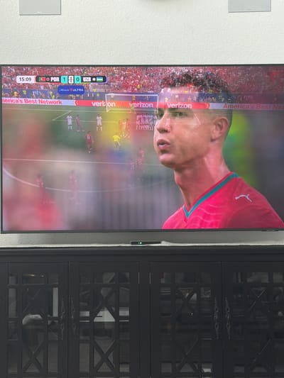 This is a close-up, slightly angled shot of a television screen displaying a soccer match. The screen is dominated by a close-up of a soccer player's face and neck, wearing a red jersey with a white Puma logo. The player appears to be sweating, suggesting an intense game. In the background, a crowd fills the stadium.
At the bottom of the screen, a scoreboard graphic indicates the time as 15:09. It shows "POR" (likely Portugal) with a score of 1 and "UZB" (likely Uzbekistan) with a score of 0. Below this, there is text including "Michelob ULTRA" and "America's Best Network". Several other logos and text elements, such as "verizon" and "FIFA", are also visible along the edges and overlays of the broadcast.
To the right of the television, a dark, ornate cabinet is partially visible. The setting appears to be indoors, with the television mounted on a wall. The lighting suggests it could be daytime, but the image is focused on the screen's content. The overall impression is of a sporting event being watched on a home entertainment system.