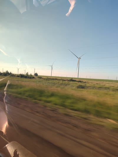 This media file depicts a rural landscape with multiple wind turbines visible across a rolling green field under a clear blue sky. The scene is captured from a moving vehicle, indicated by the motion blur in the foreground of a dirt road and the reflection of what appears to be the interior of a car on the left side of the image.
Several wind turbines are dotted across the horizon, with their tall white structures and angled blades clearly visible against the sky. Power lines stretch horizontally across the frame, connecting the turbines and adding to the impression of an energy farm. The field in the middle ground is a mix of green grass and some darker patches, suggesting vegetation or shadows. The sky is a bright, clear blue with a few faint wispy clouds. The lighting suggests it is daytime, likely late afternoon or early evening given the warm tones in the sky and the reflections.
The overall impression is one of a modern, functional landscape in a vast, open area. The motion blur in the foreground gives a sense of travel and movement. The location context provided places this scene in Майкөл ауылдық округі, Kazakhstan, suggesting a remote, possibly agricultural or steppe-like region where wind energy is being utilized. There are no visible people or text within the image. The mood conveyed is one of open space and natural, yet engineered, power generation.