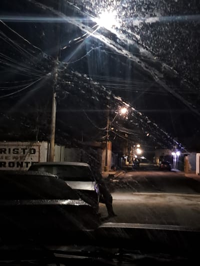 This image captures a nighttime street scene in Cumana, Venezuela, likely during or after a heavy rain. The foreground is dominated by the dark, reflective surface of what appears to be the hood of a vehicle, with streaks of water visible, suggesting recent precipitation. In the background, a street is illuminated by several streetlights, casting a glow on the wet pavement and creating reflections. Several buildings line the street, some with signs that are partially visible. One sign appears to read "CRISTO VIENE PRONTO". A utility pole with a dense network of wires stretches across the frame. A few cars are parked or driving on the street, their headlights and taillights visible. The overall atmosphere is dimly lit and damp, with a sense of urban quietude.