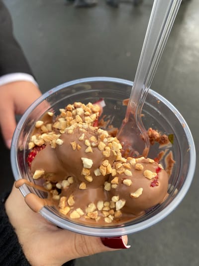 The image shows a close-up, overhead view of a clear plastic cup filled with a chocolate-covered dessert, likely strawberries, topped with chopped nuts. A clear plastic fork is partially submerged in the dessert. The dessert appears to be melting slightly, with chocolate dripping down the side of the cup. The cup is held by a person whose hand, with dark red nail polish, is visible on the right side of the frame. The background is out of focus and appears to be outdoors, possibly on a paved surface, suggesting a casual setting. The lighting indicates it is daytime, but specific weather conditions cannot be determined. There is no visible text in the image. The overall impression is of a sweet treat being enjoyed outdoors.