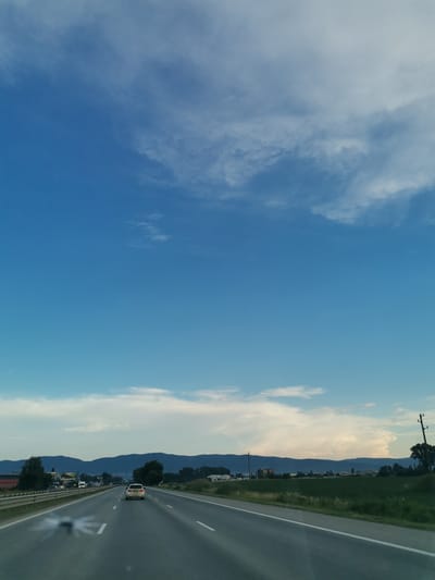 The image shows a view from a vehicle driving on a highway in Krivina, Bulgaria. The highway has multiple lanes and is bordered by a grassy median and a guardrail. In the distance, a range of mountains can be seen under a vast blue sky with scattered white clouds. The landscape includes green fields and some trees. There are other vehicles on the highway, including a white car directly ahead of the camera. Power lines and utility poles are visible along the side of the road.