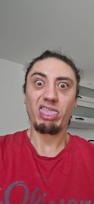 This media file features a close-up, portrait-style shot of a man making a funny face. He is wearing a red t-shirt with a large, dark blue graphic on the left side. The man has dark hair pulled back, a goatee, and is sticking his tongue out with his eyes wide open. The lighting suggests it is daytime, and the background is a plain, light-colored wall with a sliver of what appears to be a doorway or cabinet visible on the right. The man's expression conveys a playful or humorous emotion. There is some faint text visible on the t-shirt, which appears to read "Guess" or a similar word, but it is partially obscured by the fabric and the angle of the image. The scene is indoors, likely in a home environment.