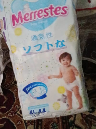 The image shows a package of Merries brand diapers. The package features a light blue and white design with pink lettering for the brand name. There is an illustration of a smiling baby wearing a diaper. Below the baby illustration, there are Japanese characters and a blue ribbon with some text inside. At the bottom of the package, there are characters "4L 44". The package is lying on a patterned carpet. The context of the image being in the city of Кызыл-Октябрь, Kyrgyzstan is not discernible from the visual information presented in the image itself.