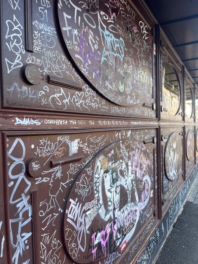 The image depicts a heavily graffitied, dark brown paneled facade in Kaiserslautern, Germany, viewed at an oblique angle from ground level. The facade features a series of rectangular panels, some incorporating large oval or circular recessed sections with decorative molding. On the right side, multiple smaller, vertically oriented rectangular panels are visible, each containing a circular glass window reflecting a blurred building across the street. An dark, narrow overhang extends above the facade.
The entire visible surface of the facade is covered extensively with multiple layers of graffiti, primarily tags and some smaller throw-ups, executed in white, light blue, purple, pink, black, and other colors. Paint drips are visible beneath some tags, notably a pink "I LOVE YOU" in the lower right. Specific legible tags include "BERND," "DAMOS," "SCHLUFFENS," "FÜNF," "NATES," "HUSSEN," "51," "TBBDOC," and "PAMC." Some Korean characters are also present. A small green circular sticker is adhered to one of the oval panels. The ground immediately adjacent to the facade is a paved sidewalk. The scene is illuminated by natural light, casting distinct shadows from the architectural elements and the overhead canopy. No people or active interactions are visible.