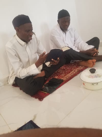 Two adult males are seated on a red patterned prayer mat on a white tiled floor in an indoor setting in Edu Town, Nigeria. Both men wear black skullcaps and white long-sleeved shirts with dark trousers. The man on the left, with his mouth slightly open, gestures with his right hand while holding an open book in his left hand. The man on the right, with a beard, holds an open book with both hands and looks towards the man on the left. Two black mobile phones lie face down on the prayer mat in front of the man on the right. To the right of the men, on the tiled floor, is a cream-colored pot with a floral design and a black lid. A small, blue patterned fabric object is partially visible on the floor in the bottom-left foreground. Both men have bare feet. The background consists of plain white walls.