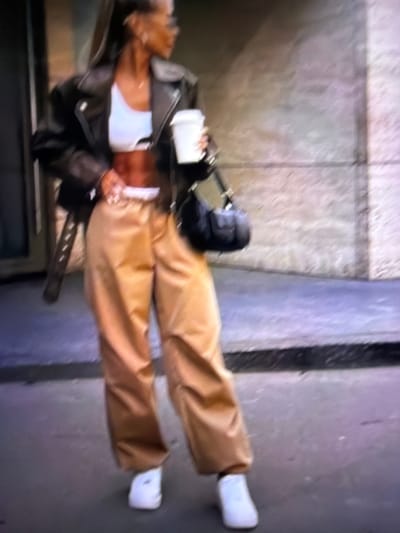 A woman stands on a sidewalk outside, holding a paper cup in her right hand and a black handbag over her left shoulder. She is wearing a black leather jacket over a white crop top, tan baggy pants, and white sneakers. The background is blurry but appears to be an urban environment with a building and a sidewalk. The specific location in the city of Tomai, Moldova, cannot be determined from the image.