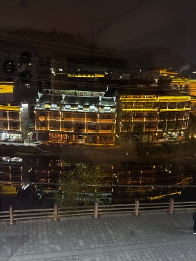 This forensic description details a nighttime scene in the city of Fenghuang, China, along what appears to be the Tuojiang River or an adjacent canal.

The scene is dominated by a row of traditional Chinese multi-story buildings, extensively illuminated with warm yellow and orange lights. These structures feature dark, gabled tiled roofs, light-colored facades, and prominent wooden balconies with intricate railings. The central building, visible as a prominent inn or guesthouse, exhibits multiple floors, with its lower levels showing brightly lit interiors, indicative of commercial establishments. Its exterior is outlined and accented by string lights and spotlights. Buildings to its immediate left and right share a similar architectural style and illumination.

Below the illuminated buildings, a dark body of water, likely a river or canal, stretches across the mid-ground. The water acts as a reflective surface, mirroring the vibrant building lights and creating elongated, distorted reflections. Sparse vegetation, including a small, leafy tree, grows along the riverbank, partially obscuring the water's edge.

In the immediate foreground, a paved stone walkway or promenade is visible, constructed from rectangular gray pavers. This walkway is bordered on the left by a low wooden fence composed of vertical posts and horizontal rails. On the far right of the walkway, a solitary individual is partially visible, walking away from the viewer. The person is wearing dark trousers or leggings and bright white shoes.

The background consists of a dark, moonless sky, with faint outlines of distant hills or mountains. Higher in the frame, several dark, parallel power lines or cables traverse the sky. The overall environment is characterized by a dense concentration of artificial light, creating a vivid nocturnal cityscape.