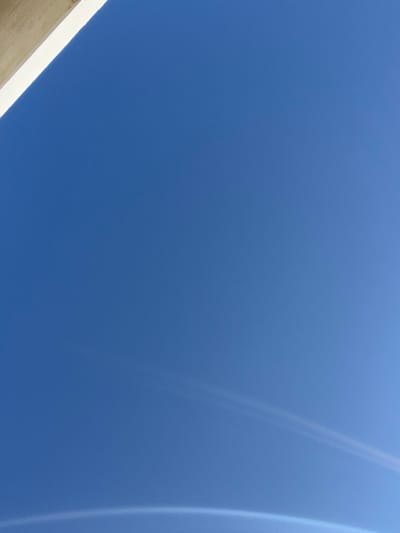 The image captures an upward view of a clear, bright blue sky, located in the city of Skagen, Denmark. No people or specific actions are depicted. In the upper left quadrant, a segment of a light-colored, likely architectural, structure is visible, featuring a white top surface and a beige underside, angled diagonally across the corner. The dominant subject is the expansive, unclouded blue sky. A single, small, dark speck, possibly an avian subject or distant aircraft, is faintly discernible in the upper-central sky. In the lower sky, two faint, parallel, curved white lines are present, suggestive of an atmospheric optical phenomenon or a highly diffused contrail. Along the bottom edge of the frame, a broad, bright, curved arc of light is evident, consistent with a sun halo component, parhelion, or significant lens flare.