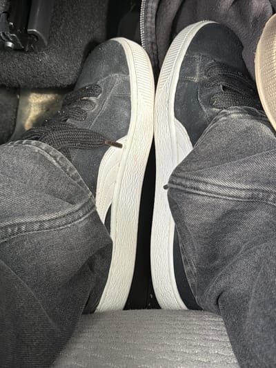 The image shows a close-up of a person's feet wearing black Puma suede sneakers and dark grey jeans. The shoes have white rubber soles and white accents on the sides. The person's feet are positioned as if they are in a car, with the floor mat and part of a car's interior visible. The perspective is from above, looking down at the feet. There is no indication of the specific location being Vallensbæk Landsby, Denmark.