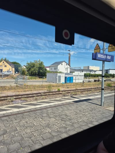The image shows a view from inside a train looking out at a train station. A blue sign with white text reads "Wittlich Hbf", indicating the station's name. There are train tracks in the foreground, a paved platform, and beyond that, buildings and vegetation. Overhead, power lines are visible against a blue sky with scattered clouds. The scene appears to be in Germany.