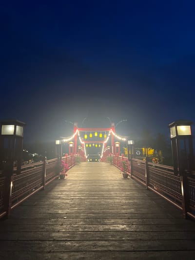 The image depicts a wooden pier at night, illuminated by decorative lights. The pier is lined with railings and lampposts, creating a pathway that recedes into the distance. Along the sides of the pier, there are planters with pink flowers. In the background, a structure with red accents and yellow lanterns is visible, suggesting an Asian architectural style. The sky is a deep blue, indicating it is nighttime. The lighting is soft, with some fog or mist adding a dreamy quality to the scene. The overall atmosphere is serene and tranquil. There is no visible text in the image. Based on the provided location context, this scene is likely in Hoi An, Vietnam.