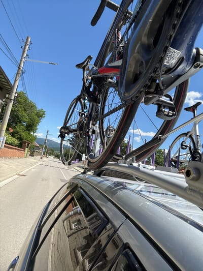 The image shows two bicycles mounted on a roof rack of a silver car. The car is driving on a paved road in Chelopech, Bulgaria. The road is lined with a sidewalk, a brick wall, and trees. In the background, there are houses and mountains. The sky is blue with some clouds. The bicycles appear to be road bikes, and one of them has a QR code sticker on its frame. The perspective is from inside the car, looking forward and slightly upwards.
