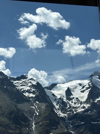 This is a photograph of the Grossglockner mountain range, near Heiligenblut am Großglockner, Austria. The image captures a dramatic vista of jagged, dark grey rocky peaks partially covered with snow and glaciers. The snowfields are broken by dark rock outcrops, and the glacial ice shows crevasses and textured surfaces. Water or meltwater channels are visible running down the snow-covered slopes.

The sky above is a vibrant blue, dotted with scattered, fluffy white cumulus clouds. A dark rectangular frame at the top of the image suggests the photograph was taken from inside a vehicle or building, possibly through a window, as there are some faint reflections visible on the surface. The overall impression is one of a majestic and rugged alpine landscape under a clear, bright sky.