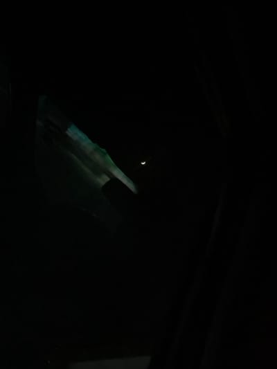A sliver of a crescent moon is visible in the upper left corner of the frame, against an otherwise dark sky. The foreground is dominated by the interior of a vehicle, seen at night. The light from the moon reflects subtly on the dashboard and windshield, giving a vague suggestion of texture and shape. The car's ceiling is barely visible at the top of the image.