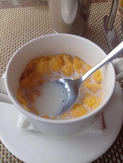 A white mug sits on a white saucer, filled with cornflakes and milk. A silver spoon rests inside, partially submerged in the milk. A crumpled white napkin cushions the mug. The saucer and mug are on a textured woven mat. A silver teapot is partially visible in the background, along with a silver knife. The light reflects off the spoon and teapot, creating highlights.