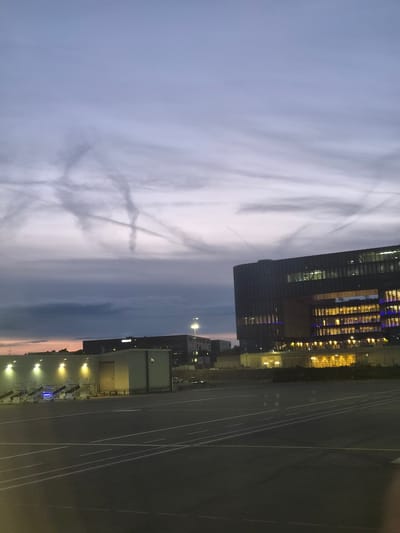 This image captures a nighttime scene in Niederanven, Luxembourg. The sky, predominantly grey and pale blue, showcases faint contrails forming an "X" shape. A dark, modern building with illuminated windows stands adjacent to a large, dark paved area marked with white lines. A smaller, light-colored building with yellow lighting is also visible. Overall, the environment appears urban and lit by artificial light sources.