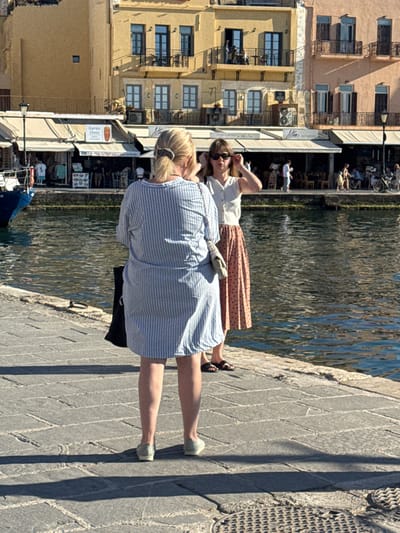 In the city of Chania, Greece, two women are standing on a cobblestone walkway adjacent to a body of water. The woman in the foreground, viewed from the back, wears a blue and white striped dress and light-colored shoes. She carries a black bag. The woman in the background, facing forward, wears a white top and a polka-dot skirt. She is adjusting her sunglasses. Behind them, a row of buildings with a mix of yellow and peach facades line the waterfront. Shops with awnings are visible at street level, and there are people seated at outdoor tables. The water reflects the sunlight and the surrounding structures.