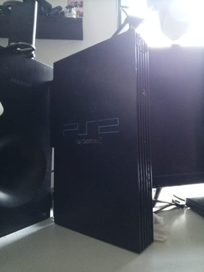 The scene depicts an indoor arrangement of electronic equipment on a light-colored surface in the city of Villanueva de Gállego, Spain. A black Sony PlayStation 2 Slim console stands vertically in the foreground, displaying the "PS2" logo and "PlayStation 2" text. A small red indicator light is visible on its upper right edge. To the left of the console, a black speaker or subwoofer, partially branded with "SAMSUNG," is visible. To the right, a dark monitor or television screen is present. A bright, diffused light source, possibly a window, is visible behind the monitor, casting a strong glare. In the upper left background, a dark, wall-mounted shelf holds several indistinct items, including what appears to be a glass jar and other small objects. No individuals are present, and no specific actions or interactions are observed.