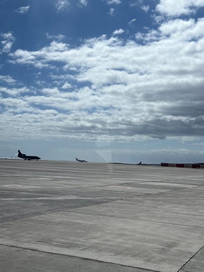 The image captures an airport tarmac under a partly cloudy sky, located within the city of Granadilla de Abona, Spain, consistent with Tenerife South Airport. The foreground and midground are dominated by a vast expanse of light grey concrete airfield pavement, featuring discernible expansion joints and faint, circular ground markings. In the left midground, a dark-colored, possibly black, corporate jet with rear-mounted engines and a T-tail is visible, appearing stationary or slowly taxiing left. To its right, a smaller, light-colored twin-engine aircraft, likely a turboprop or small jet, is also present on the tarmac, seemingly stationary or in slow motion. In the distant right background, a row of red and white striped barriers is visible, behind which a section of a dark red aircraft is partially obscured. The sky overhead features a mix of clear blue patches and varying formations of white and grey stratocumulus clouds, denser on the right. No individuals are visible. The scene is viewed from a slightly elevated perspective, possibly through a window, suggested by a subtle reflection.