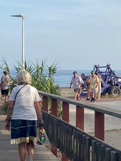 This media file depicts a scene on a sunny day, likely in Resinera-voladilla, Spain. The foreground features a person from behind, wearing a white t-shirt and blue shorts, holding a hat. In the midground, a group of people are gathered near a blue vehicle, possibly a tractor or a trailer, on a paved area next to a sandy beach and the ocean. The people appear to be casually dressed, in shorts and t-shirts, suggesting a relaxed, outdoor setting. The presence of the vehicle could indicate some sort of work or activity related to the beach or coastal area. The lighting suggests it is daytime. The overall atmosphere seems casual and warm, consistent with a beachside location. There is no visible text in the image.
