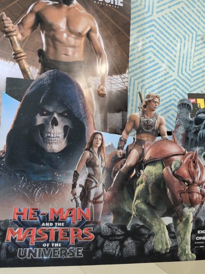 This image is a poster for "He-Man and the Masters of the Universe." In the foreground, a muscular man with blonde hair, wearing a chest plate and brown shorts, rides a large, green, feline-like creature with a red, helmet-like adornment on its head. The creature has sharp claws and yellow eyes. To the left of the rider, a woman in a white and brown outfit is visible, holding a sword. Behind them, a large, hooded figure with a skeletal face and glowing red eyes looms. At the bottom of the poster, the title "HE-MAN AND THE MASTERS OF THE UNIVERSE" is displayed in large, red, 3D lettering. In the upper right corner, a portion of a blue and white patterned background is visible, along with a section of a castle and a "get" logo, suggesting this is a promotional display. The poster is placed on a wall, and the lower portion of the image shows a floor. The poster appears to be located at the GVK One Mall in Hyderabad, India, as indicated by promotional materials and the context of such posters often being displayed in malls.