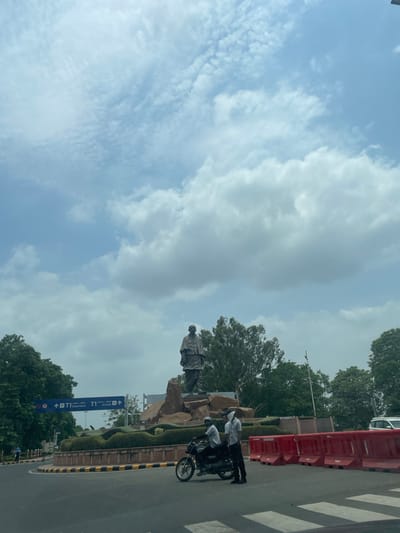 The image depicts the Statue of Unity, a colossal statue located in Gujarat, India, which is situated near the Sardar Sarovar Dam on the Narmada river, in the Kevadiya Colony, in the state of Gujarat, India. In the foreground, two men on a motorcycle are observed, one driving and the other seated behind. The motorcycle is positioned on a road with a pedestrian crossing. Behind them, red traffic barriers are visible. The statue itself is situated in the background, atop a rocky base. The sky above is filled with clouds. A traffic sign indicating "T1 Departures" and "T1 Arrivals" is also visible, suggesting proximity to an airport. Trees and landscaping surround the statue and the road.