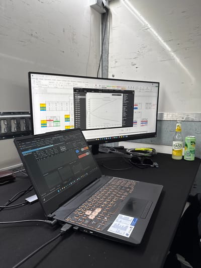 The scene is inside a white truck, potentially a race team's support vehicle, located in Stavelot, Belgium (latitude: 50.442, longitude: 5.97). A black work surface is visible, covered in various equipment and cables. Two computer screens display data. The left screen shows a data table and the right screen displays a graph titled "Tork Setting Versus Engine Speed." A black laptop is open on the work surface, with its keyboard illuminated. A black leather wallet is visible on the left side of the work surface, while two bottles of drink, one glass and one aluminum can, are situated on the right side of the surface. There are also black headphones and a small USB drive on the table. The space appears to be a mobile work area. The scene is devoid of people.