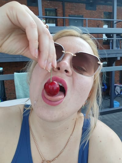 In Pirogovo, Russia, a close-up, waist-up, outdoor shot captures a woman with blonde hair and light skin wearing sunglasses and a blue swimsuit. She is holding a single cherry with her right hand and extending it towards her open mouth, with her tongue visible. The background shows a brick building with balconies and railings. The lighting suggests it is daytime.