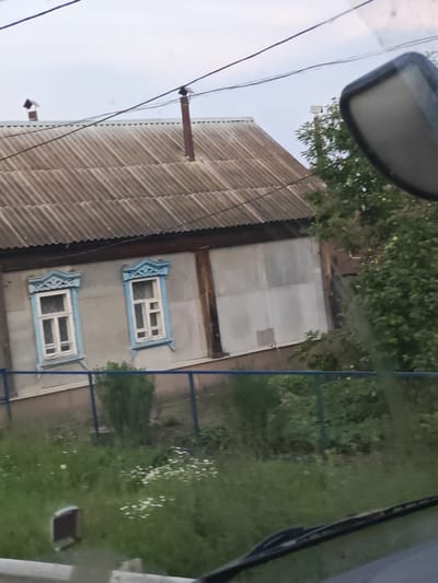 The image shows a rural house with a corrugated metal roof, two windows with blue decorative frames, and what appears to be a wooden structure on the side. A blue fence runs in front of the house, with overgrown grass and some white wildflowers in the foreground. A portion of a car's windshield and a side mirror are visible in the foreground, suggesting the photo was taken from a vehicle. The sky is overcast. The location is identified as Makhylino, Russia.