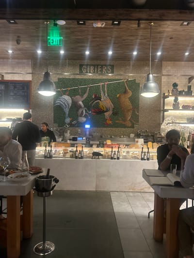 A restaurant scene presents a mosaic mural of game birds behind a cheese and wine display. Patrons sit at tables. One table is in the foreground. An "exit" sign is visible. Overhead lighting fixtures hang from the ceiling.