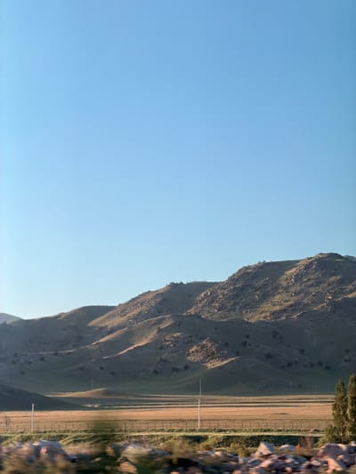 A landscape view in Chüy, Kyrgyzstan shows a mountain range adjacent to a flat field with scattered vegetation and power lines. The left portion of the image is dominated by a clear, light blue sky. A portion of some rocks can be seen on the right.