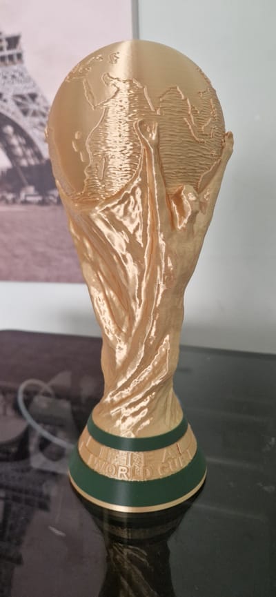 The image shows a close-up, rotated view of a golden trophy, resembling the FIFA World Cup. The trophy has a base with text that reads "FIFA WORLD CUP". The globe portion of the trophy is detailed, depicting continents. The trophy is presented against a blurred background that includes what appears to be images of the Eiffel Tower. The lighting suggests an indoor setting, possibly a display or exhibition. The overall impression is of a prestigious award. The location context provided is South Holland, United Kingdom, though there are no explicit visual cues in the image to confirm this specific location. There are no people visible in the image, and no specific weather conditions can be inferred.