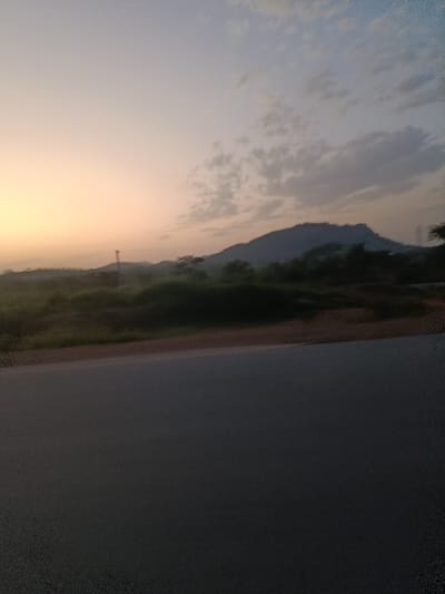 The image depicts an outdoor landscape in Jere North, Nigeria, under low light conditions, indicative of either sunrise or sunset. The lower third of the frame is dominated by a dark grey asphalt road surface. Immediately adjacent to the road, a narrow strip of reddish-brown earth or gravel is visible, followed by an expanse of dense, low-lying green and dark brown vegetation. In the left-midground, a tall, slender, dark-silhouetted structure, consistent with a utility pole or antenna, extends upwards. The background features a large, dark mountain or hill range silhouetted against the horizon, partially obscured by atmospheric haze. The sky occupies the upper half of the image, displaying a gradient from a light orange-yellow hue on the left to a pale blue-grey on the right, with scattered, indistinct clouds distributed throughout. No individuals, animals, or explicit activities are present in the scene.
