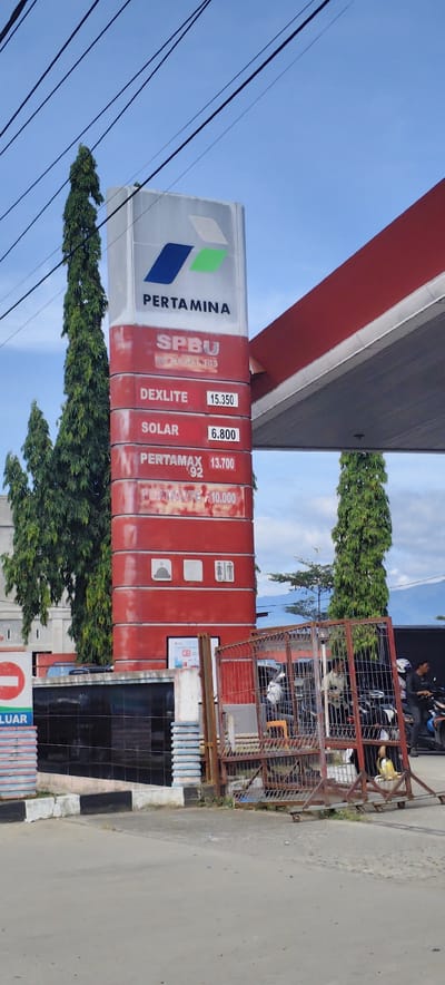 The image depicts a Pertamina gas station in Mali Guyui, Indonesia. A tall sign displays the Pertamina logo and fuel prices: Dexlite at 15,350, Solar at 6,800, Pertamax 92 at 13,700, and Pertalite at 10,000. The sign also includes restroom icons. A red gas station canopy is visible to the right of the sign. In the foreground, a weathered metal barrier stands near a black tiled structure. Several individuals are present, some on motorcycles, near the station. Trees and power lines are also visible in the background.