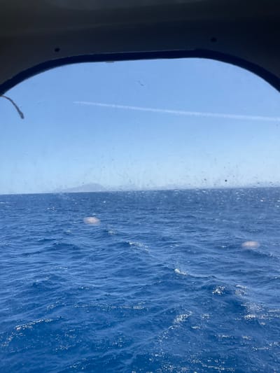 The image displays an expansive view of the open sea and sky, observed through an arched window. The foreground features deep blue, moderately agitated ocean water, illuminated by direct sunlight creating numerous shimmering reflections on its surface. A low, hazy landmass, possibly distant islands or coastline, is visible along the horizon. The sky above is clear and light blue, traversed by a single, prominent white aircraft contrail positioned horizontally across the upper third of the frame. The windowpane itself exhibits several small, diffuse light artifacts and minor surface imperfections. This maritime scene is observed from the sea in the vicinity of Marsala, Italy. No human subjects, specific vessels, or additional objects are identifiable within the primary view beyond the natural elements and the viewing portal.