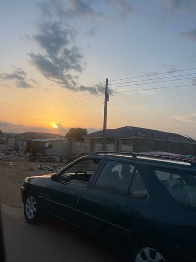 The image depicts an outdoor scene at sunset or sunrise in Rinjim Gani, Nigeria, characterized by warm light from a low sun. The sky transitions from a bright orange-yellow near the horizon, where the sun is partially obscured by clouds, to lighter blues and grays at the top, with scattered cumulus clouds present.
In the foreground, a dark-colored Peugeot 306 Break (station wagon) is positioned on the right, with its passenger side visible. The vehicle appears to be on a paved or hardened dirt surface. A light-colored item, possibly a blanket or animal, is discernible through the rear passenger window. Reflections of the sun are visible on the car's bodywork.
The midground features a dirt road or open ground on the left, strewn with significant amounts of litter, including plastic bags and debris. Several single-story buildings are visible, some constructed with corrugated metal sheets, others with concrete walls and corrugated roofs. One concrete wall includes what appear to be handwritten phone numbers or codes: "0803713006" and "08092758022." This wall is topped with barbed wire fencing.
Multiple human figures are present in the midground, near the structures and litter. One figure, possibly a child, stands on the far left near the corrugated metal structures. Another figure is seated or crouched amidst the litter closer to the center. Their specific actions are indistinct due to distance and lighting.
A wooden utility pole with multiple horizontal power lines extends vertically through the mid-left portion of the frame, carrying lines across the sky. In the background, beyond the immediate structures, additional indistinct buildings and sparse vegetation are visible under the horizon line.