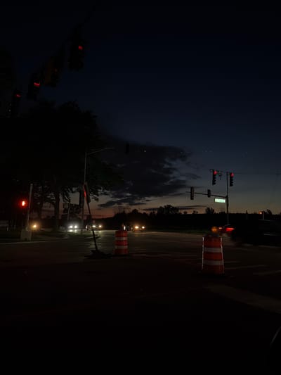 The image depicts a street intersection at dusk in Buffalo Grove, Illinois, United States of America. Traffic lights are visible, with some displaying red signals. Several orange and white construction barrels are placed on the roadway. In the background, trees line the horizon, and clouds are present in the darkening sky. Headlights of cars are visible in the distance, indicating ongoing traffic. A street sign with green lettering on a white background is also present.