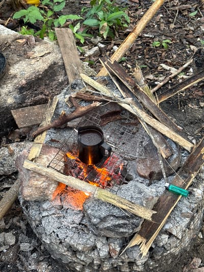 A black metal cup sits on a wire grill over a fire. The fire burns within a rock structure covered in wire mesh. Broken sticks and twigs are scattered around the fire pit. A green lighter rests on a rock next to the fire. Green plants grow to the left of the scene. The location is Moo 7, Thailand.