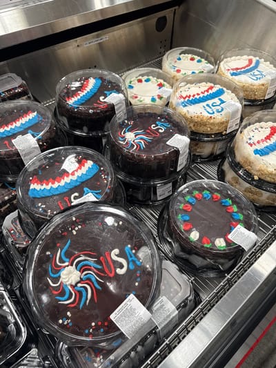 In the city of Yonkers, United States of America, a display of approximately ten round cakes sits within a refrigerated case. The cakes are decorated with patriotic red, white, and blue frosting, many featuring the word "USA." Some cakes are chocolate with chocolate frosting, while others are white cake with white frosting and crushed nut exteriors. The cakes are packaged in clear plastic containers and are stacked on metal shelves. A sign with the text "SAFE LOAD LIMIT" is visible above the cakes. The overall impression is a commercial setting, likely a bakery or grocery store, presenting holiday-themed desserts.