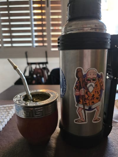 This image captures an indoor scene in Villa San Andrés, Argentina, featuring a traditional mate setup. In the foreground, a brown calabash mate gourd with a polished silver-colored metal rim, decorated with a repeating geometric pattern, sits on a dark, flat surface. The gourd contains green yerba mate leaves, with a silver bombilla (straw) inserted. To the immediate right, a tall, cylindrical stainless steel thermos stands upright. This thermos has a black plastic base and an integrated black handle/pour mechanism on its right side; its upper section consists of a clear, possibly glass or plastic, liner. A partially obscured circular blue sticker is visible on the left side of the thermos, and a prominent sticker depicting an animated elderly male character with a white beard, sunglasses, an orange Hawaiian-style shirt, shorts, and flip-flops, holding a staff, is affixed to the right side of the thermos. A white, knitted or crocheted mat is partially visible beneath the mate gourd on the left. The background is blurred, showing a window with white horizontal louvered blinds through which sunlight filters, creating distinct horizontal strips of light and shadow. Indistinct dark shapes of furniture or other objects are visible further in the background.