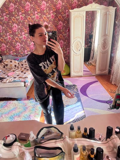 A young woman is taking a mirror selfie in a bedroom. She is wearing a black t-shirt with a graphic print and black leggings. The room is decorated with floral wallpaper and has a large, ornate mirrored wardrobe. A purple and abstract patterned rug covers part of the floor, and a fluffy, rainbow-colored rug is also visible. On a surface to the right, there is a collection of makeup and beauty products, including several bottles and compacts. The lighting suggests it is daytime. The woman's expression is neutral. The location is identified as Krasnoe-na-Volge, Russia.