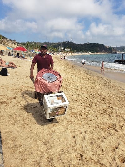 A man stands on a sandy beach next to a box-like vendor cart with a round metal top covered by pink fabric. He wears a maroon polo shirt and dark shorts. The beach scene includes people in the distance near the ocean and several umbrellas. The sky is partly cloudy. The image is horizontally oriented.