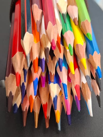 This is a close-up, eye-level shot of a pile of colored pencils. The pencils are arranged diagonally from the top left to the bottom right of the frame. The tips of the pencils are sharpened and some are in use, showing a slight bit of wear. The colors visible include red, purple, blue, orange, yellow, green, pink, brown, and black. The pencils are tightly packed, creating a textured and layered effect. The background is dark and appears to be a textured surface, possibly a desk or fabric. The lighting is even, suggesting an indoor setting. There are no people or animals visible in the image, and no text is present. The overall impression is one of art supplies ready for use, or perhaps a jumble of pencils after a creative session. The context of Nalchik, Russia, does not provide any specific visual cues within the image itself, as the subject matter is universal.