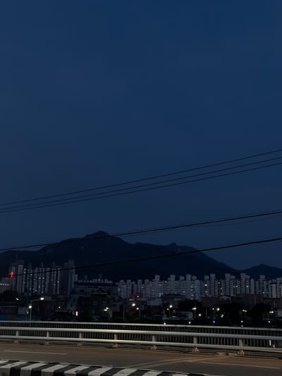 The image shows a view of Uijeongbu-si, South Korea, at dusk. The foreground features a bridge with a metal railing and a road marked with yellow and black stripes. Beyond the bridge, a cityscape unfolds with numerous apartment buildings and some smaller structures. Streetlights are visible, illuminating parts of the urban landscape. In the background, dark, silhouetted mountains rise against the deep blue twilight sky. Power lines are strung across the upper portion of the image, adding a linear element to the composition. No people are clearly visible in the image. The overall scene depicts a city transition from day to night, with the urban environment set against a natural mountainous backdrop.