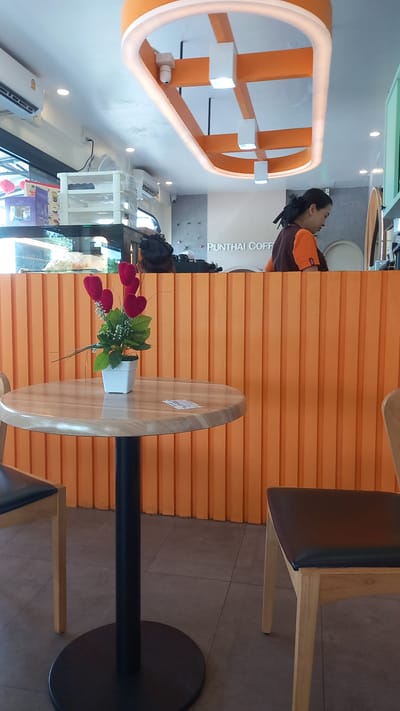 The image shows an interior scene in a café called Punthai Coffee in Plai Phraya, Thailand. A woman wearing an orange and brown uniform is partially visible behind an orange ribbed wall. A round wooden table with a black pedestal is present, decorated with a small vase of artificial red tulips and greenery. Next to the table, a chair with a wooden frame and black seat is also visible. A decorative orange oval-shaped lighting fixture is visible in the background. Outside the window, greenery can be seen.
