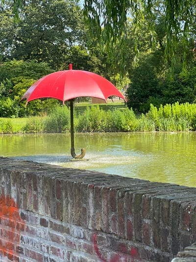 The image shows a red umbrella sculpture in a pond. The sculpture is a fountain, with water spraying out from beneath the umbrella. The pond is surrounded by lush green trees and vegetation. In the foreground, there is a brick wall with some graffiti on it. The image appears to be taken on a sunny day, and the water in the pond is a bright green color, reflecting the surrounding foliage and the sky. The overall scene is serene and picturesque. This sculpture is located in the Prinsentuin (Prince's Garden) in Leeuwarden, Netherlands.