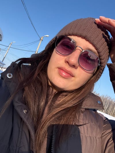 A woman wearing a brown beanie and sunglasses looks up at the camera. She is wearing a brown puffer jacket. Clear blue sky is visible in the background along with trees and a snow-covered ground. Electrical poles and wires are also visible against the blue sky.