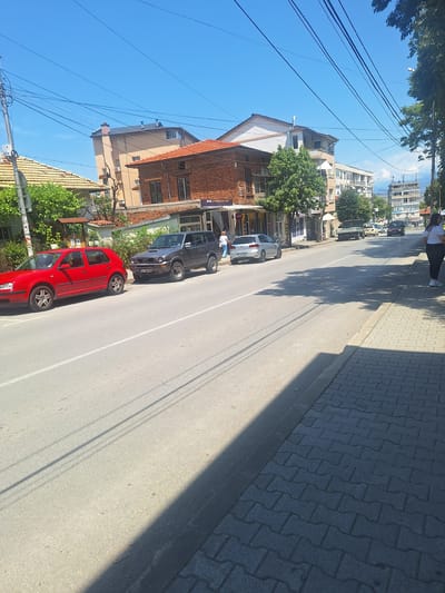 The image displays a sunny street scene in Petrich, Bulgaria, likely captured during mid-morning or late afternoon due to the long, distinct shadows cast by trees and buildings across the road and sidewalk. The street runs down the center of the frame, lined on both sides by multi-story buildings.

On the left side of the asphalt road, two cars are prominently parked: a red four-door Volkswagen Golf (Mk4) in the foreground, and behind it, a dark-colored SUV with a visible Bulgarian license plate "E 1530 KA," which confirms the Blagoevgrad region where Petrich is located. Further down the street, another light-colored vehicle is parked. The road features a clear white dividing line.

To the right, a wide sidewalk is paved with dark, intricately patterned "bone-shaped" interlocking pavers. Tree foliage is visible, and the shadows of trees and utility poles stretch across the pavement and road. Several people are present: one individual in a white top and dark pants stands on the sidewalk in the upper right, while another person in a blue shirt is near the dark SUV.

The buildings along the street exhibit varied architecture, with some featuring red tiled roofs and brick facades, while others have lighter plaster exteriors. Ground-floor establishments appear to be commercial spaces. Visible text on shop signs includes "ЗАКУСКИ" (Snacks/Breakfast) and "СБОРНИ ТУРИСТИЧЕСКИ ПЪТЕШЕСТВИЯ АГЕНЦИЯ" (Group Tourist Trips Agency), indicating local businesses. A dense network of black utility wires crisscrosses against a clear, bright blue sky overhead. The overall scene captures a typical, vibrant urban environment on a clear day.