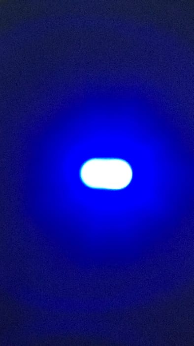 The image displays a bright, horizontally elongated white light source positioned centrally within a dark blue, grainy field. A blue luminescence emanates from the light source, diffusing into the surrounding dark blue background. No discernible architectural features, landmarks, individuals, or specific environmental identifiers are present to determine a specific location within Lompoc, United States, or any other city. The scene is devoid of additional subjects, objects, actions, or interactions.
